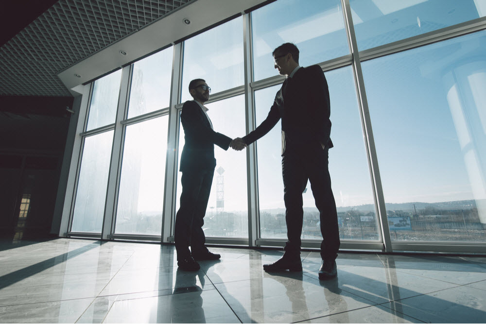 Two businessmen are shaking hands with each other standing against panoramic windows