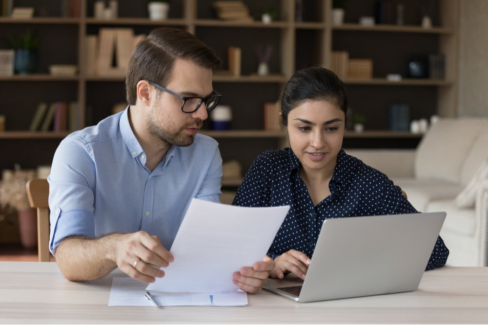 Two diverse coworker employees sharing laptop, discussing paper report at workplace